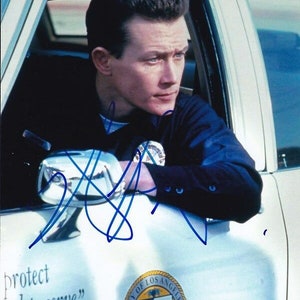 May include: A man in a blue uniform sits in a white car. He is looking to the right. The car has the words "protect and to serve" and the seal of the City of Los Angeles on the side. The man's signature is visible on the car.