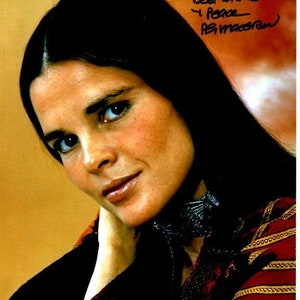 Może przedstawiać: Portret zbliżeniowy kobiety z długimi, ciemnymi włosami. Ma na sobie czerwoną koszulę w złote wzory i czarny szal. Kobieta patrzy w obiektyw z delikatnym uśmiechem. Obraz jest podpisany "Best wishes & Peace - Ali MacGraw".