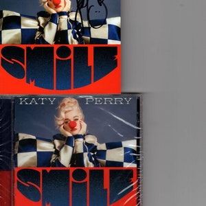 Puede incluir: Una caja de CD con un patrón de cuadros rojos y azules. La caja presenta una foto de Katy Perry con un disfraz de payaso con una nariz roja. El texto "KATY PERRY" y "SMILE" está impreso en la caja.