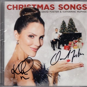 May include: A CD cover featuring a woman holding a snow globe with a man playing a piano inside. The snow globe is surrounded by presents. The cover is signed by David Foster and Katharine McPhee. The title of the album is "Christmas Songs" by David Foster & Katharine McPhee.