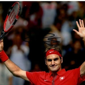 May include: A man in a red shirt with a Swiss flag on the chest raises his right hand in the air while holding a tennis racket. He is wearing a black headband with a white logo.