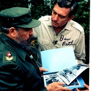 Pode incluir: Um homem com uniforme militar verde e um boné verde com uma estrela está olhando para um álbum de fotos com outro homem. O homem com o uniforme militar tem barba. O outro homem está vestindo uma camisa marrom clara e assinou seu nome "Dan Rather" no álbum.
