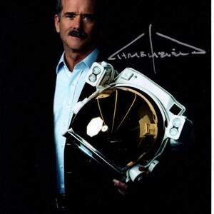 Puede incluir: Un hombre con traje y camisa azul sostiene un casco de astronauta blanco con una visera dorada. El casco tiene una firma en el lateral que dice "Jim Lovell".