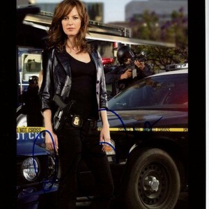 May include: A woman wearing a black leather jacket and black trousers stands in front of a police car. She is holding a gun in her right hand. The car is a black and white police car with yellow caution tape around it. The text on the tape reads "Police Line Do Not Cross".
