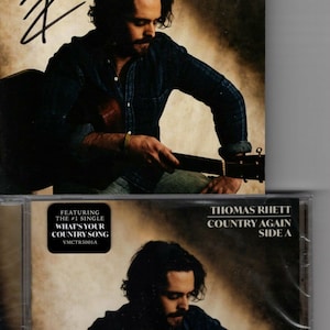 Puede incluir: Una caja de CD con una foto de Thomas Rhett sosteniendo una guitarra. La caja es negra con un borde blanco y el texto "Thomas Rhett Country Again Side A" está impreso en la parte delantera. La caja de CD presenta el single número 1 "What's Your Country Song" y el número de álbum VMCTR5001A.