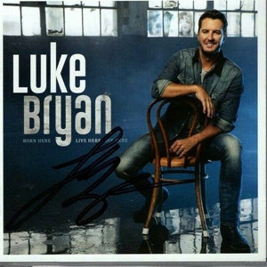 Puede incluir: Una portada de CD con el cantante de música country Luke Bryan. La portada es azul con texto blanco que dice "Luke Bryan" y "Born Here Live Here Die Here". Bryan está sentado en una silla de madera con una camisa vaquera azul y pantalones vaqueros. Está mirando a la cámara y sonriendo.