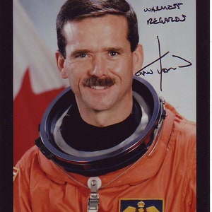 Puede incluir: Un retrato de cerca de un hombre que lleva un mono naranja con cuello negro y un parche circular en el pecho. El parche tiene el texto "Chris Hadfield" y una bandera canadiense. El hombre tiene bigote y sonríe. El texto "with my warmest regards" y una firma están escritos con tinta azul en la esquina superior derecha de la imagen.