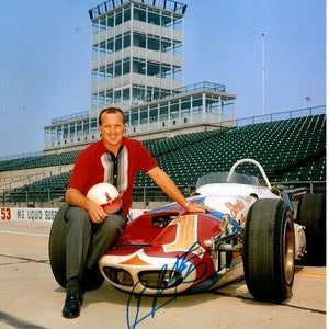 May include: A man in a red and white striped shirt sits next to a white, red, and blue race car. He is holding a white helmet with a red number one on it. The car has a number one on the side and the words "Springfield" and "Liquid Suspensions" on it. The car is on a racetrack with a tower in the background.