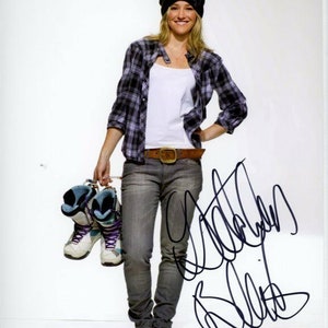 Puede incluir: Una mujer con una camisa de cuadros blanco y negro, un top blanco sin mangas, pantalones vaqueros azules y una boina negra. Lleva un par de botas de esquí azules y blancas. La mujer sonríe y tiene el pelo rubio. El fondo es blanco. La firma de la mujer está en tinta negra debajo de ella.