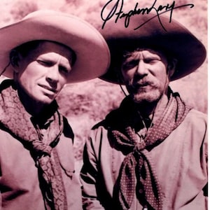 May include: Two men wearing cowboy hats and bandanas pose for a photo. The man on the left is wearing a light brown hat and a dark brown bandana. The man on the right is wearing a dark brown hat and a light brown bandana. The man on the right has a signature that reads "Stephen Lang" above his head.