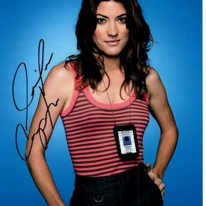 May include: A signed photograph featuring a person wearing a pink and black striped tank top and dark pants. A badge hangs around the neck. The background is a solid blue. The signature is in black ink.