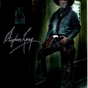 May include: A black and white photo of a man wearing a cowboy hat, a blue shirt, and jeans. He is holding a gun in his right hand. The photo is signed "Stephen Lang".