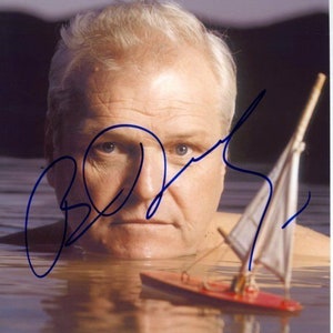 May include: A man with white hair is submerged in water up to his neck. He is looking at the camera. A small toy sailboat is floating in the water in front of him. The man's signature is written in blue ink on the image.