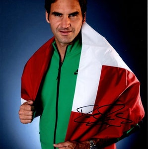 May include: A man wearing a green jacket with a white and red flag draped over his shoulders. The flag has a white stripe in the middle and red stripes on either side. The man is smiling and looking at the camera. He is wearing a watch on his left wrist. The flag is signed with a black signature.