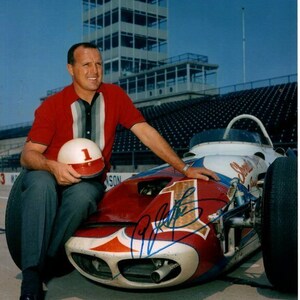 May include: A man in a red and white striped shirt sits next to a white and red race car. He is holding a white helmet with a red number one on it. The car has a large number one on the side and the driver's signature.