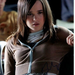 May include: A young woman with long brown hair wearing a brown zip-up sweater with a blue zipper and a white shirt underneath. She is holding a pen in her hand and looking down.