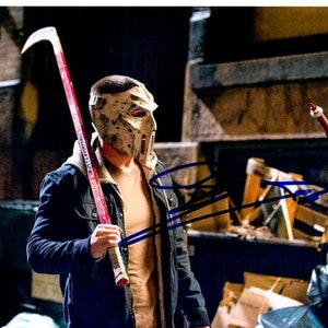 May include: A man wearing a white and black hockey mask holds a red hockey stick. He is wearing a dark blue jacket over a tan shirt. The background is blurry and appears to be an urban setting.