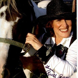 May include: A woman wearing a black cowboy hat and a white shirt with embroidery is petting a black and white horse. The woman is smiling and has signed the photo "Love, Ruth Bussie".