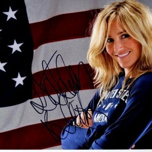 Puede incluir: Una persona sonriente con cabello rubio lleva una camiseta azul de manga larga con el texto blanco "UNITED STATES OF AMERICA". El fondo muestra la bandera estadounidense y una firma.