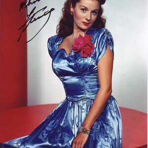 Peut inclure: Une femme vêtue d'une robe en satin bleu à motif floral et d'une fleur rouge sur le corsage. Elle porte un bracelet au poignet et ses cheveux sont coiffés en boucles. L'image est signée "Rhonda Fleming".
