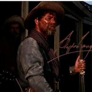 May include: A man wearing a brown cowboy hat and a plaid shirt is holding a knife. He is standing in front of a building with a wooden door. The man's signature, "Stephen Lang", is written in cursive in the upper right corner of the image.
