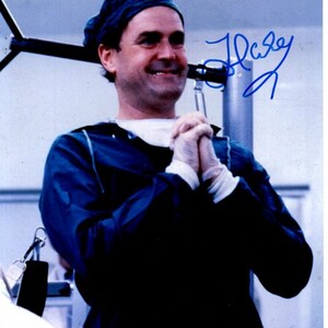 May include: A man wearing a blue surgical cap and scrubs smiles and claps his hands together. The image is signed "Halsey".