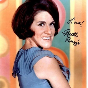 May include: A woman with short brown hair wearing a blue sleeveless shirt smiles at the camera. The background is a colorful abstract design. The woman's autograph is in black ink and reads "Love! Ruth Buzzi".