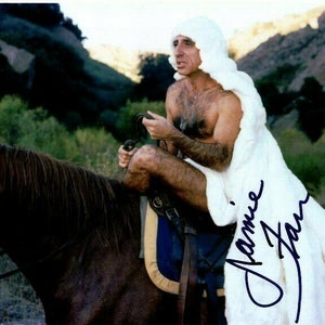 May include: A man wearing a white robe rides a brown horse. The man is bare-chested and has a white wig on. The robe has the signature "Jamie Farr" written in blue ink.