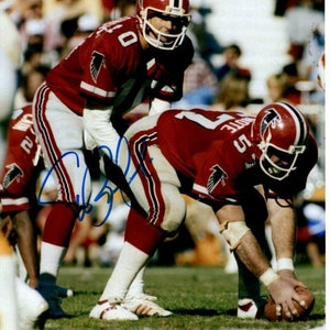 Può includere: Due giocatori di football americano con uniformi rosse e bianche con il logo degli Atlanta Falcons sui loro caschi e maglie. Il giocatore in primo piano sta tenendo un pallone da football ed è in posizione di calcio. Il giocatore sullo sfondo è in piedi con le mani sui fianchi.