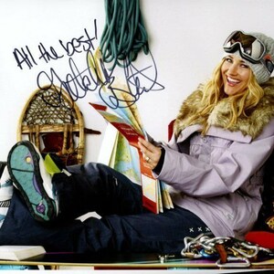 Puede incluir: Una mujer con una chaqueta morada con cuello de piel y gafas de esquí está sentada en un banco con un mapa en la mano. Ella está sosteniendo un mapa y tiene una firma que dice "All the best!"