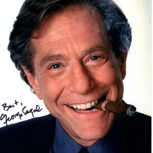 May include: A man with short brown hair is smiling and holding a lit cigar in his mouth. He is wearing a blue shirt and a dark suit jacket. The man is looking at the camera and has a friendly expression. The background is white.