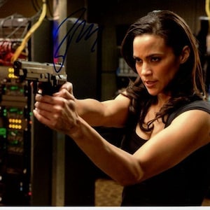 May include: A woman in a black shirt holds a silver handgun in her right hand. She is looking directly at the camera with a serious expression. The background is a blurry image of a server rack.