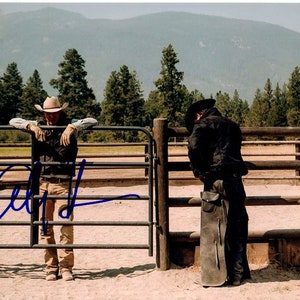 Könnte beinhalten: Zwei Cowboys stehen an einem Holzzaun. Ein Cowboy trägt einen braunen Cowboyhut und eine braune Weste. Der andere Cowboy trägt einen schwarzen Hut und eine schwarze Jacke. Der Cowboy mit dem braunen Hut hält ein Seil. Der Hintergrund ist ein Feld mit Bäumen und Bergen.