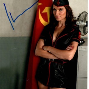 May include: A woman wearing a black leather outfit with red trim and a red star on her hat. She is standing in front of a red flag with a yellow hammer and sickle.