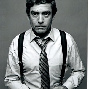 Pode incluir: Uma fotografia a preto e branco de um homem a usar uma camisa branca, uma gravata de riscas e suspensórios. Ele está a segurar uma chávena branca nas suas mãos. O homem está a olhar para a câmara com uma expressão séria. A fotografia está assinada "Dan Rather".