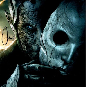 May include: A close-up image of a person with blue eyes and a textured face, holding a gray mask. The person's face is partially visible, with a detailed, textured appearance. The mask is smooth and has a single large eye hole.