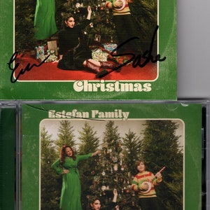 Puede incluir: Una portada de álbum de Navidad verde y blanca con una familia de cuatro personas de pie frente a un árbol de Navidad. La portada del álbum tiene el texto "Estefan Family" y "Christmas". La portada del álbum está firmada por los miembros de la familia.