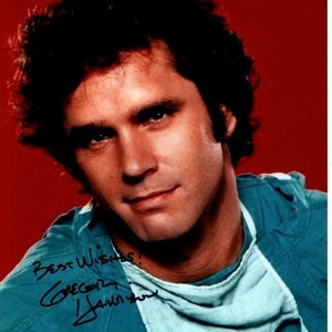 May include: A close-up of a man with dark hair wearing a blue surgical scrub top. He is looking at the camera and smiling. The man has signed his name in black ink on the scrub top, "Best Wishes! Gregory Harrison".