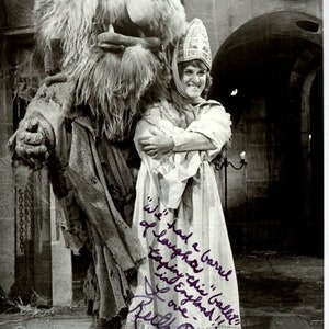 May include: A black and white photo of a woman in a white dress and a crown, holding a furry monster. The woman is smiling and the monster is looking at the camera. The photo is signed by Ruth Buzzi with the text "We had a barrel of laughter doing this ballet! Love - England!!"
