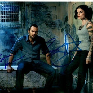 Puede incluir: Un hombre y una mujer están de pie en una habitación con una bañera blanca. El hombre lleva una camisa azul y pantalones vaqueros. La mujer lleva un top gris sin mangas y pantalones negros. Las paredes están cubiertas de grafitis.