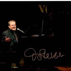 May include: A man in a black suit and tie is singing into a microphone in front of a black piano. The signature "Don Reber" is written in gold cursive on the black surface in front of the piano.
