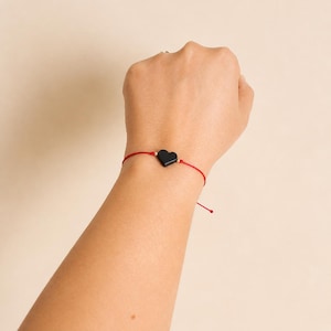 May include: A red string bracelet with a black heart-shaped charm. The bracelet is tied around a wrist. The charm is centered on the red string. The background is a solid, neutral color.