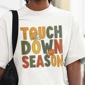 Op de afbeelding: Een wit t-shirt met de tekst "TOUCH DOWN SEASON" in een retro lettertype. Het ontwerp bevat een footballhelm en een football. De persoon draagt een spijkerbroek en een zwarte schoudertas.