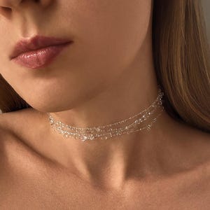 May include: A delicate, layered choker necklace with clear, sparkling beads. The necklace is made of multiple strands of tiny, transparent beads, creating a shimmering effect. The jewelry is worn around the neck.