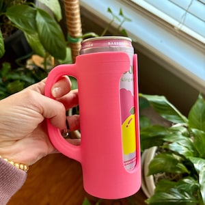 May include: A bright pink can cooler with a handle, holding a can of sparkling energy drink. The can is visible through a cutout on the cooler, displaying the brand name and flavor. The cooler is held in a hand.
