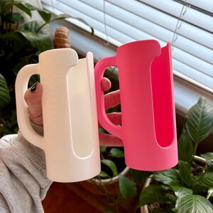 May include: Two mug-shaped drink holders, one white and one bright pink, are held up against a background of green plants and a window. Each holder has a handle and a slot to hold a beverage container.