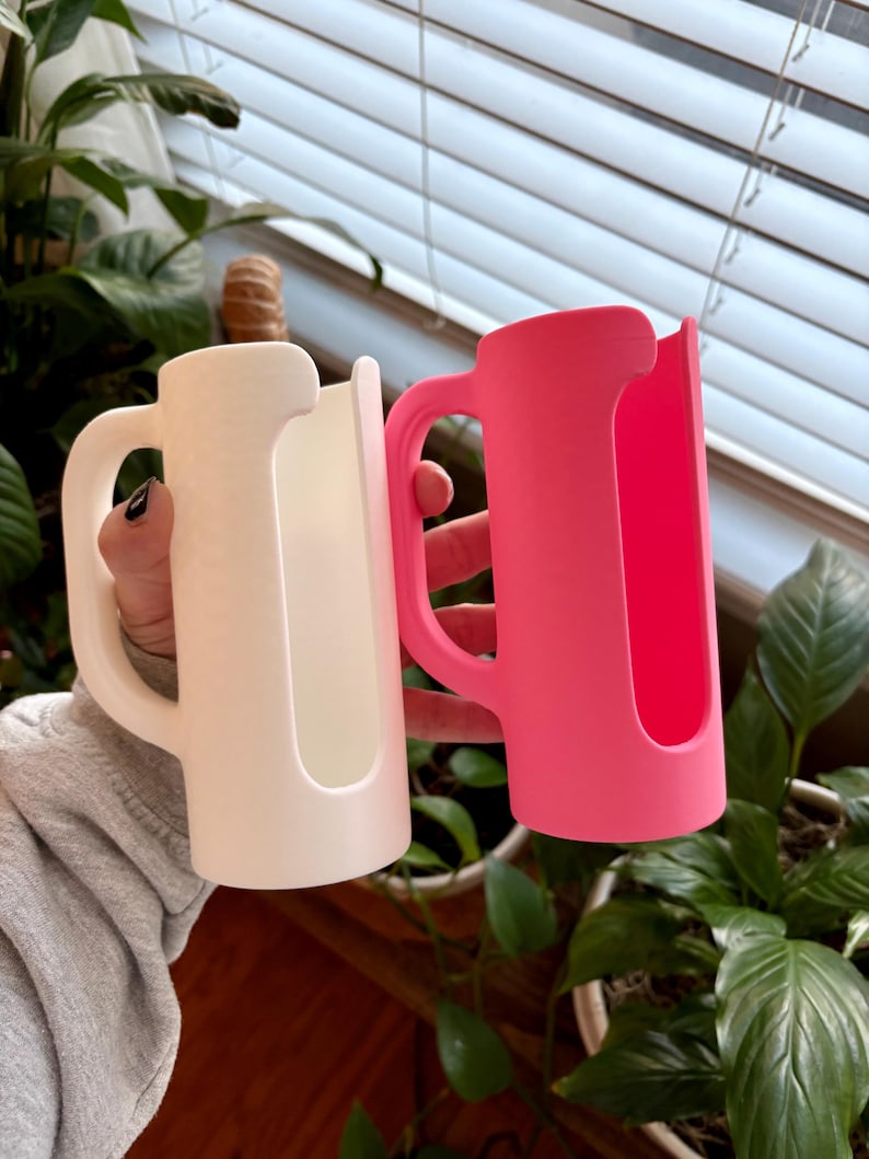 May include: Two mug-shaped drink holders, one white and one bright pink, are held up against a background of green plants and a window. Each holder has a handle and a slot to hold a beverage container.