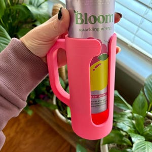 May include: A pink can holder with a handle, holding a can of Bloom sparkling energy drink. The can is light purple with the words "Raspberry Lekce" and "Bloom sparkling energy" printed on it. The holder is made of a flexible material.