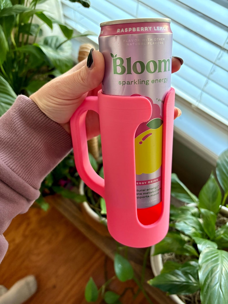 May include: A pink can holder with a handle, holding a can of Bloom sparkling energy drink. The can is light purple with the words "Raspberry Lekce" and "Bloom sparkling energy" printed on it. The holder is made of a flexible material.