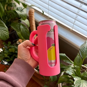 May include: A bright pink can cooler with a handle, holding a can of raspberry lemon sparkling drink. The can is visible through a cutout in the cooler. The cooler is held in front of a background of green plants.
